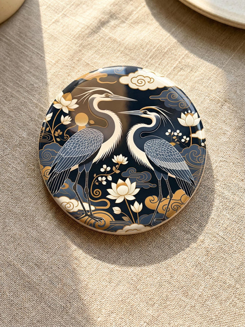 A round 4-inch drink coaster by Grimoire and Goblet. The coaster features a highly reflective glossy top surface displaying two Great Blue Herons, lotus flowers, and stylized clouds in metallic antique gold and slate blue against a solid, deep navy blue background. The edges reveal a natural brown, porous cork backing underneath. The intricate design resembles vintage Chinoiserie and botanical natural history art, perfectly suited for protecting tables from beverages.