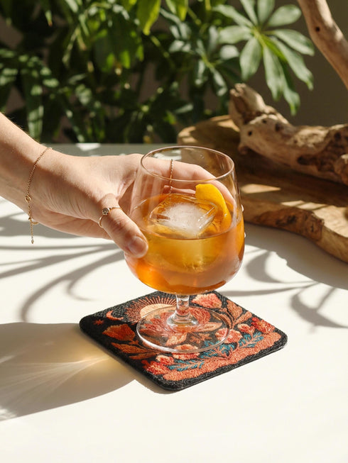 A Grimoire and Goblet drink coaster featuring a rustic faux burlap texture print. The coaster displays a gothic botanical design in dark charcoal ink on a tan jute-like polyester background. The smooth fabric surface mimics a woven look. Ideal for Dark Academia decor and protecting tables from spills. The design includes a central motif with vintage styling.