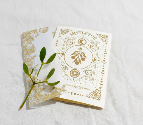 A Grimoire & Goblet luxury Mistletoe ritual card set. The 5x7 cream greeting card features gold celestial designs framing “Mistletoe” and “Love, Protection & New Beginnings.” Shown with a vellum wrap printed in gold florals, a white wax seal with mistletoe, and a premium envelope. Includes a 4x6 ritual insert on handmade deckled paper titled “Ritual for Peace and Protection.” A spiritual Yule stationery set designed as a luxurious unboxing experience.