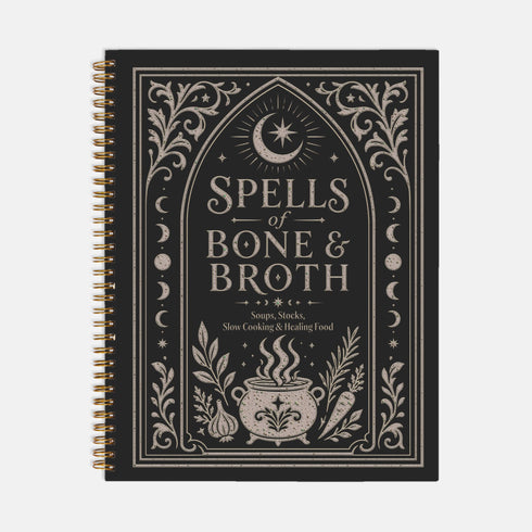 Spiral bound hardcover recipe journal lying flat. The matte black front cover is illustrated with light beige botanical vines, small crescent moons, a radial starburst, and a central cauldron releasing curls of steam, with classical lettering reading Spells of Bone and Broth. A black metal wire-o coil binds the left edge. The interior shows bright white pages printed with a structured recipe template, including labeled sections for ingredients and instructions, over a rigid chipboard backing.