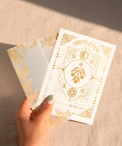 A Grimoire & Goblet luxury Mistletoe ritual card set. The 5x7 cream greeting card features gold celestial designs framing “Mistletoe” and “Love, Protection & New Beginnings.” Shown with a vellum wrap printed in gold florals, a white wax seal with mistletoe, and a premium envelope. Includes a 4x6 ritual insert on handmade deckled paper titled “Ritual for Peace and Protection.” A spiritual Yule stationery set designed as a luxurious unboxing experience.