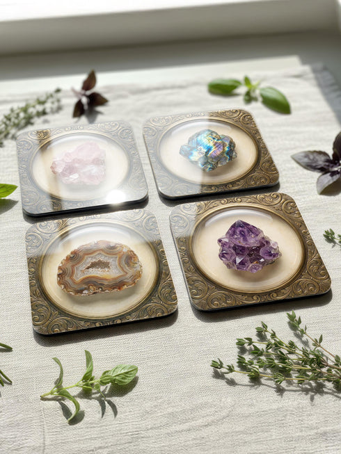 Grimoire and Goblet set of four square glossy coasters featuring high-resolution printed illustrations of raw crystals: purple amethyst, pink rose quartz, iridescent labradorite, and banded agate. Each gemstone is depicted inside a printed faux antique brass baroque frame with intricate scrollwork, set against a cream parchment background. The design is a flat 2D trompe l'œil print on hardboard, creating a realistic 3D specimen box illusion.