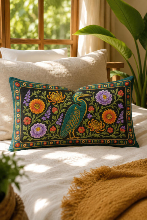 Heron lumbar pillow cover featuring a teal wading bird with gold detailing standing among orange marigolds, golden chrysanthemums, purple peonies, lavender sprigs, and coral red blooms with green foliage on a deep green black panel. A teal border with gold rope trim and a folk style band of red rosettes frames the design. Rectangular 20 x 12 inch cushion case with hidden zipper, the same tapestry look design printed on both sides in smooth, linen feel polyester fabric.