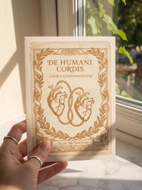 A vintage-style greeting card titled "DE HUMANI CORDIS" featuring a medical illustration of two anatomical hearts connected by a single looped blood vessel. The hearts are drawn in a sepia and crimson woodcut style on a cream background mimicking aged parchment. Text below reads "Fig. 14: The Twin Pulse." The card evokes a dark academia or cottagecore aesthetic. Inside text reads "Two different beats, one shared rhythm.