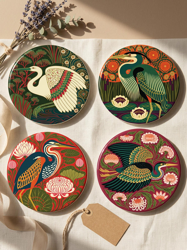 This set of four round ceramic coasters features a glossy, smooth, high-fidelity flat-printed surface. Each piece displays highly detailed Art Nouveau style illustrations of herons and egrets surrounded by stylized water lilies, chrysanthemums, and rich foliage. The vibrant, intricate color palette includes deep emerald greens, rich crimson reds, navy blues, and golden yellows. The sides are smooth, unprinted white ceramic, and the underside is fitted with a natural brown cork backing.