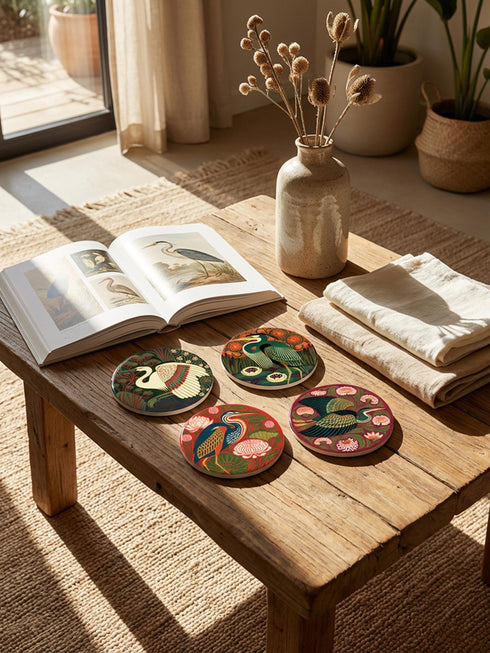 This set of four round ceramic coasters features a glossy, smooth, high-fidelity flat-printed surface. Each piece displays highly detailed Art Nouveau style illustrations of herons and egrets surrounded by stylized water lilies, chrysanthemums, and rich foliage. The vibrant, intricate color palette includes deep emerald greens, rich crimson reds, navy blues, and golden yellows. The sides are smooth, unprinted white ceramic, and the underside is fitted with a natural brown cork backing.