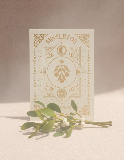 A Grimoire & Goblet luxury Mistletoe ritual card set. The 5x7 cream greeting card features gold celestial designs framing “Mistletoe” and “Love, Protection & New Beginnings.” Shown with a vellum wrap printed in gold florals, a white wax seal with mistletoe, and a premium envelope. Includes a 4x6 ritual insert on handmade deckled paper titled “Ritual for Peace and Protection.” A spiritual Yule stationery set designed as a luxurious unboxing experience.