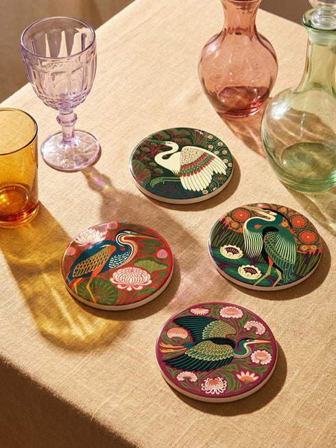 This set of four round ceramic coasters features a glossy, smooth, high-fidelity flat-printed surface. Each piece displays highly detailed Art Nouveau style illustrations of herons and egrets surrounded by stylized water lilies, chrysanthemums, and rich foliage. The vibrant, intricate color palette includes deep emerald greens, rich crimson reds, navy blues, and golden yellows. The sides are smooth, unprinted white ceramic, and the underside is fitted with a natural brown cork backing.