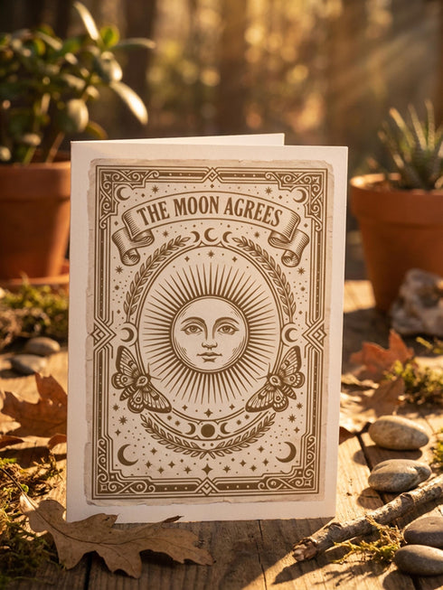 Grimoire and Goblet greeting card featuring a vintage woodcut style illustration of a radial sun with a serene human face. The sun is flanked by two Luna moths and sits above a Triple Moon symbol. A banner above reads THE MOON AGREES in gothic font. Printed in burnished gold tones on cream paper. Interior text reads Yours in this lifetime and the next. Dark Academia and Whimsigoth aesthetic stationery 5x7 card with matching envelope for gothic romance or altar decor.