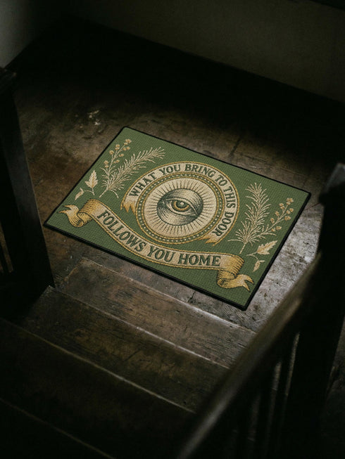 A rectangular doormat featuring a printed faux embroidery design on a muted olive green background. The symmetrical artwork centers on an all-seeing eye inside a circular medallion with a radiating sunburst pattern. An antique gold banner curves above reading WHAT YOU BRING TO THIS DOOR. A matching banner below reads FOLLOWS YOU HOME. The central eye is flanked by detailed botanical fern fronds and delicate wildflowers. The mat is finished with a sleek black fabric border trim.