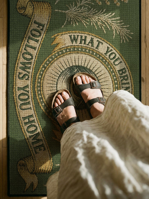 A rectangular doormat featuring a printed faux embroidery design on a muted olive green background. The symmetrical artwork centers on an all-seeing eye inside a circular medallion with a radiating sunburst pattern. An antique gold banner curves above reading WHAT YOU BRING TO THIS DOOR. A matching banner below reads FOLLOWS YOU HOME. The central eye is flanked by detailed botanical fern fronds and delicate wildflowers. The mat is finished with a sleek black fabric border trim.