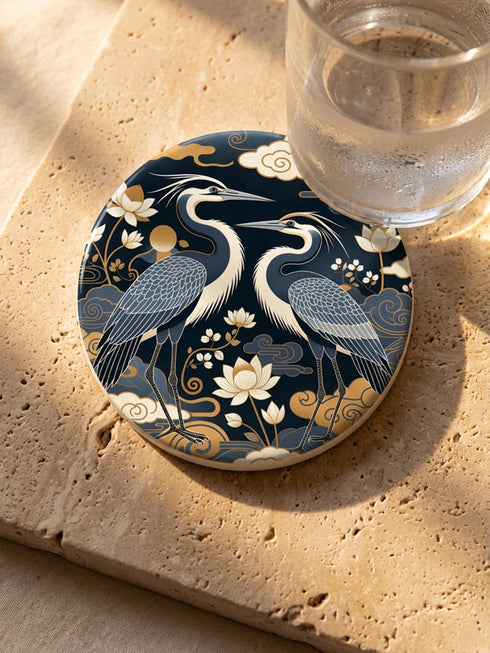 A round 4-inch drink coaster by Grimoire and Goblet. The coaster features a highly reflective glossy top surface displaying two Great Blue Herons, lotus flowers, and stylized clouds in metallic antique gold and slate blue against a solid, deep navy blue background. The edges reveal a natural brown, porous cork backing underneath. The intricate design resembles vintage Chinoiserie and botanical natural history art, perfectly suited for protecting tables from beverages.