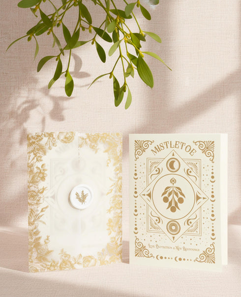 A Grimoire & Goblet luxury Mistletoe ritual card set. The 5x7 cream greeting card features gold celestial designs framing “Mistletoe” and “Love, Protection & New Beginnings.” Shown with a vellum wrap printed in gold florals, a white wax seal with mistletoe, and a premium envelope. Includes a 4x6 ritual insert on handmade deckled paper titled “Ritual for Peace and Protection.” A spiritual Yule stationery set designed as a luxurious unboxing experience.