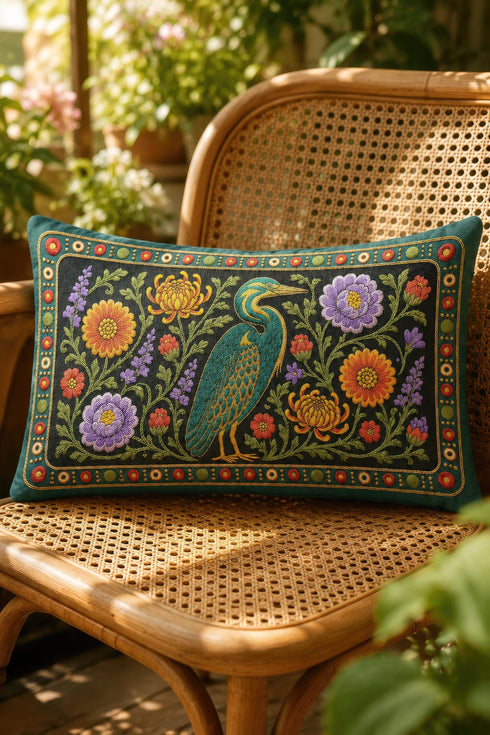 Heron lumbar pillow cover featuring a teal wading bird with gold detailing standing among orange marigolds, golden chrysanthemums, purple peonies, lavender sprigs, and coral red blooms with green foliage on a deep green black panel. A teal border with gold rope trim and a folk style band of red rosettes frames the design. Rectangular 20 x 12 inch cushion case with hidden zipper, the same tapestry look design printed on both sides in smooth, linen feel polyester fabric.