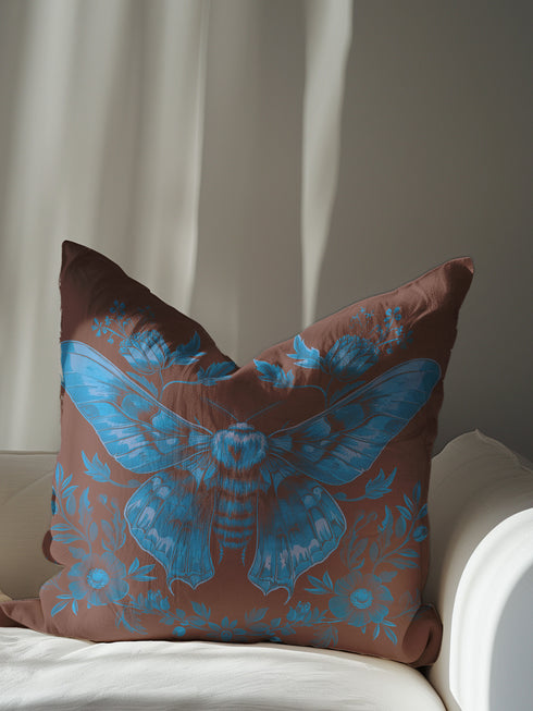 A decorative pillow cover featuring a large Luna moth illustration in electric blue on a rich chocolate brown background, adorned with symmetrical botanical elements and flowers in a detailed vintage naturalist style.
