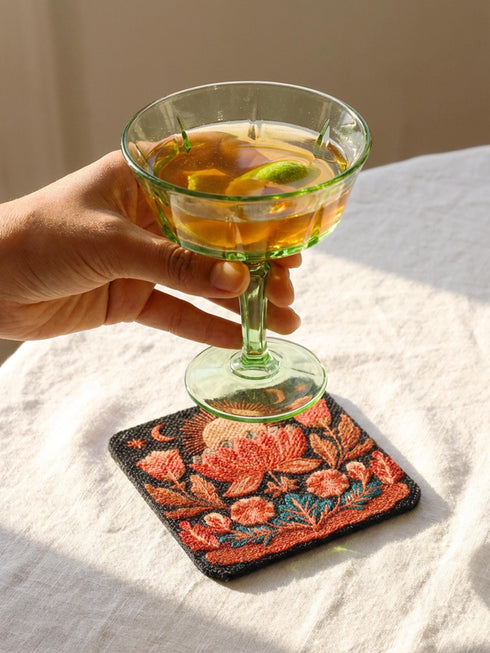 A Grimoire and Goblet drink coaster featuring a rustic faux burlap texture print. The coaster displays a gothic botanical design in dark charcoal ink on a tan jute-like polyester background. The smooth fabric surface mimics a woven look. Ideal for Dark Academia decor and protecting tables from spills. The design includes a central motif with vintage styling.