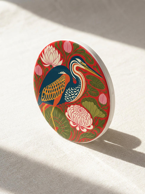 This set of four round ceramic coasters features a glossy, smooth, high-fidelity flat-printed surface. Each piece displays highly detailed Art Nouveau style illustrations of herons and egrets surrounded by stylized water lilies, chrysanthemums, and rich foliage. The vibrant, intricate color palette includes deep emerald greens, rich crimson reds, navy blues, and golden yellows. The sides are smooth, unprinted white ceramic, and the underside is fitted with a natural brown cork backing.