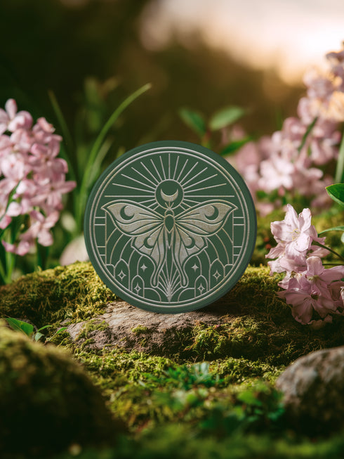 A round, dark hunter green ceramic drink coaster with a non-slip cork back. The top surface features an intricate, engraved-style celestial moth design in a light beige or off-white color. The luna moth is centered under a crescent moon and a radiant sunburst. The design is symmetrical and includes stars and geometric line art, evoking a gothic, witchy, and spiritual aesthetic. The coaster has a matte finish and is designed as a piece of functional art.