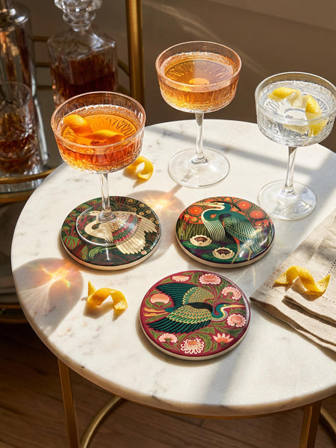This set of four round ceramic coasters features a glossy, smooth, high-fidelity flat-printed surface. Each piece displays highly detailed Art Nouveau style illustrations of herons and egrets surrounded by stylized water lilies, chrysanthemums, and rich foliage. The vibrant, intricate color palette includes deep emerald greens, rich crimson reds, navy blues, and golden yellows. The sides are smooth, unprinted white ceramic, and the underside is fitted with a natural brown cork backing.