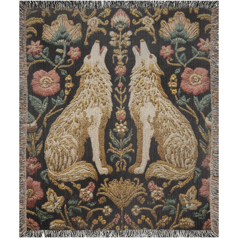 A rectangular, 100% woven cotton throw blanket with a detailed jacquard design. The throw features a symmetrical, vertical pattern of two howling wolves in profile, rendered in shades of beige, gold, and brown against a solid black background. The wolves are surrounded by intricate floral and foliage motifs. The thick, textured weave is visible. The blanket is finished on all four sides with a two-inch, multi-colored fringe that includes black, white, red, and gold threads.