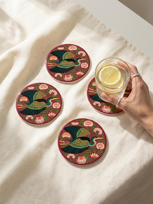 A round, 4-inch ceramic drink coaster featuring a glossy, flat-printed Art Nouveau design. The central motif is a flying heron with teal plumage and scalloped golden wing feathers, surrounded by stylized winding olive green foliage, peach chrysanthemums, and botanical blooms. The artwork is set against a solid, deep blood-red background. The coaster has a smooth white edge and a natural brown cork backing. Available as a single coaster or in matching sets of four or six.