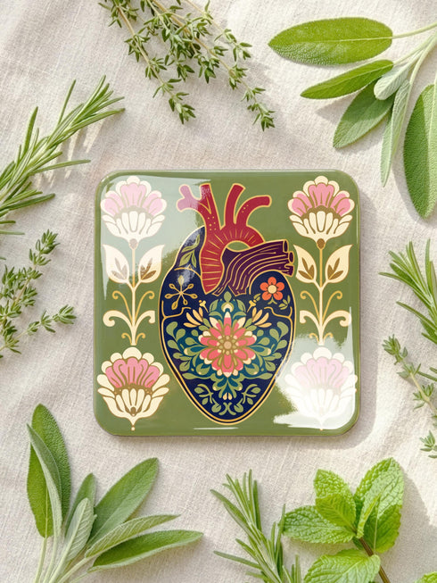 A set of four premium 4x4 inch anatomical heart coasters from Grimoire and Goblet. Each square coaster is made of masonite hardboard with a high-gloss finish and a non-slip cork base. The designs feature intricate biological heart illustrations with folk-art floral motifs in vibrant crimson, pink, yellow, and purple against mossy green and deep teal backgrounds. The high-fidelity print mimics a luxury embroidered texture, perfect for a cardiology gift or dark academia home decor.