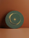 Round ceramic coaster with forest green background. Golden crescent moon embraces a sun at the center. Fine golden lines radiate outward, creating a sunburst pattern to the edge. White rim indicates cork backing. Coaster diameter is 4.04 inches.