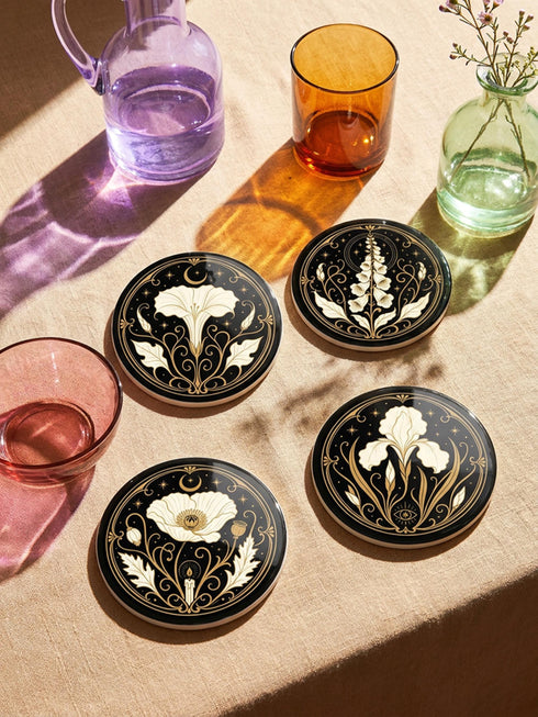 A set of four round 4-inch ceramic coasters featuring a glossy 2D flat-printed Art Deco botanical design. Each coaster displays a different cream-colored flower including a moonflower, foxglove, poppy, and iris, set against a solid black background. The floral motifs are framed by intricate gold linework, crescent moons, four-pointed stars, and mystical symbols like a candle and an all-seeing eye. The coasters have a smooth white edge and are finished with a natural brown cork backing.
