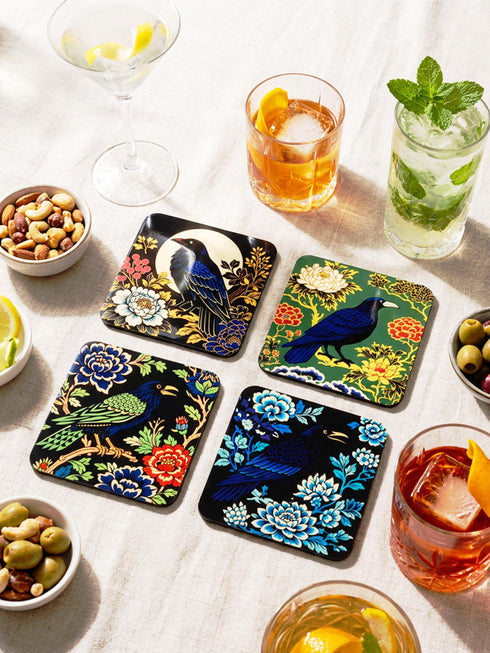 Grimoire and Goblet set of 4 glossy square coasters featuring Chinoiserie style raven illustrations. 1: Navy blue with blue floral pattern and raven. 2: Navy background with black raven, white full moon, and red berries. 3: Moss green background with blue bird and yellow chrysanthemums. 4: Black background with teal-winged raven and red peonies. MDF hardboard with cork backing. Flat vector botanical art style.