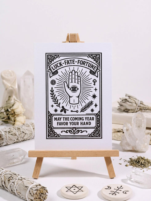 Grimoire and Goblet presents a 5x7 inch pearlescent greeting card. The design features a black linocut-style illustration of an open palm with an eye, surrounded by rays, keys, coins, and stars. The cover reads Luck Fate Fortune and May The Coming Year Favor Your Hand. The interior features a smaller hand icon and a printed blessing. The paper has a luxury silvery shimmer finish.