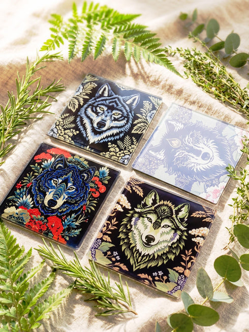 Set of 4 square tempered glass coasters featuring a mystical wolf design on a black background. High-gloss finish with non-slip rubber feet. Dark Academia and Whimsigoth aesthetic home decor. Close-up detail shows grey wolf fur texture and amber eyes. Durable drinkware protection for wooden tables. Grimoire and Goblet gothic luxury barware accessory for coffee tables and desks. Heat resistant glass surface with polished edges.