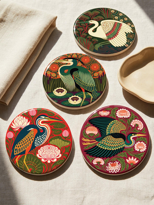 This set of four round ceramic coasters features a glossy, smooth, high-fidelity flat-printed surface. Each piece displays highly detailed Art Nouveau style illustrations of herons and egrets surrounded by stylized water lilies, chrysanthemums, and rich foliage. The vibrant, intricate color palette includes deep emerald greens, rich crimson reds, navy blues, and golden yellows. The sides are smooth, unprinted white ceramic, and the underside is fitted with a natural brown cork backing.