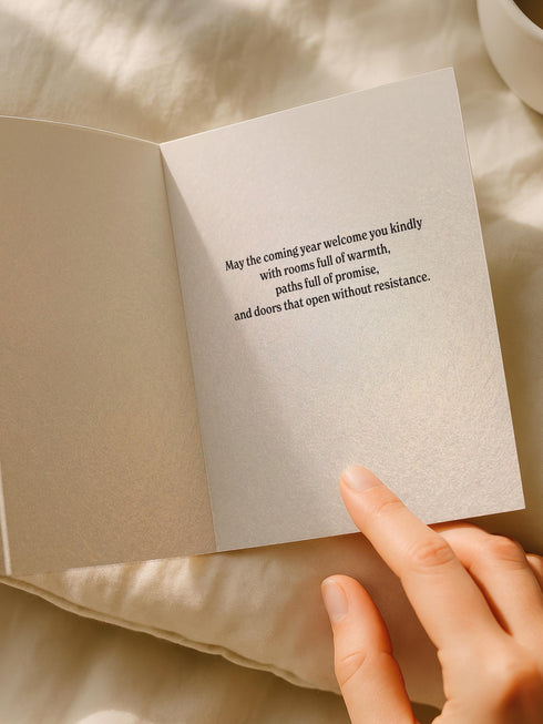 A vertical 5x7 inch pearlescent greeting card by Grimoire and Goblet featuring a high-contrast black woodcut-style illustration. The design centers on a gothic cathedral archway framing a glowing candle and a winding path leading forward. Surrounding the arch are intricate floral vines and stars. A banner at the top reads A YEAR OF OPEN DOORS in serif typography. The paper has a subtle silvery shimmer. The inside contains a printed poem about warmth and promise. Perfect for spiritual gifting.