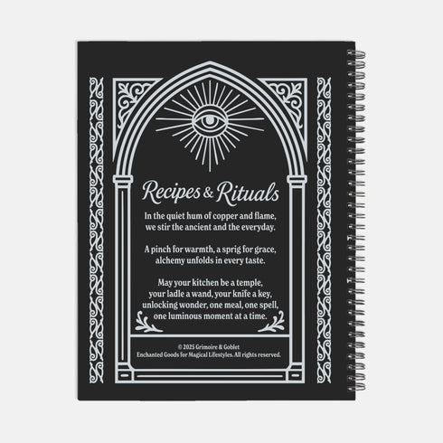 A black spiral-bound hardcover recipe journal lies flat. The cover is matte black with a white line-art design titled "Recipes & Rituals." The art nouveau style illustration features an all-seeing eye under a sunburst, framed by an ornate arch. Below the eye, a crossed spoon and knife are centered above a mortar and pestle filled with herbs. The entire design is bordered by intricate botanical flourishes. The notebook is 8.5 x 11 inches with a wire-o binding, a lay-flat cookbook for kitchen use.
