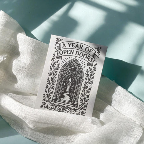 A vertical 5x7 inch pearlescent greeting card by Grimoire and Goblet featuring a high-contrast black woodcut-style illustration. The design centers on a gothic cathedral archway framing a glowing candle and a winding path leading forward. Surrounding the arch are intricate floral vines and stars. A banner at the top reads A YEAR OF OPEN DOORS in serif typography. The paper has a subtle silvery shimmer. The inside contains a printed poem about warmth and promise. Perfect for spiritual gifting.