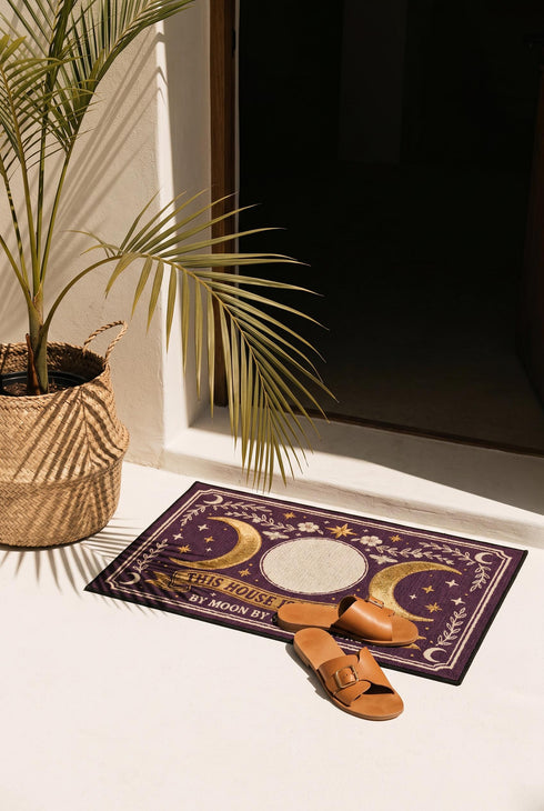 A rectangular doormat featuring a printed faux embroidery design on a deep plum purple background. The symmetrical artwork centers on a large cream full moon flanked by two golden crescent moons, creating a Triple Moon symbol. Above the moons are gold stars, cream flowers, and two small bees. Below, a golden scroll banner reads "THIS HOUSE IS PROTECTED" with "BY MOON BY STAR BY BONE" underneath. The mat is framed by cream leafy vines and finished with a black fabric border trim.