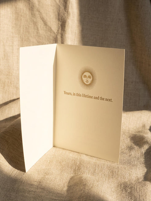 Grimoire and Goblet greeting card featuring a vintage woodcut style illustration of a radial sun with a serene human face. The sun is flanked by two Luna moths and sits above a Triple Moon symbol. A banner above reads THE MOON AGREES in gothic font. Printed in burnished gold tones on cream paper. Interior text reads Yours in this lifetime and the next. Dark Academia and Whimsigoth aesthetic stationery 5x7 card with matching envelope for gothic romance or altar decor.