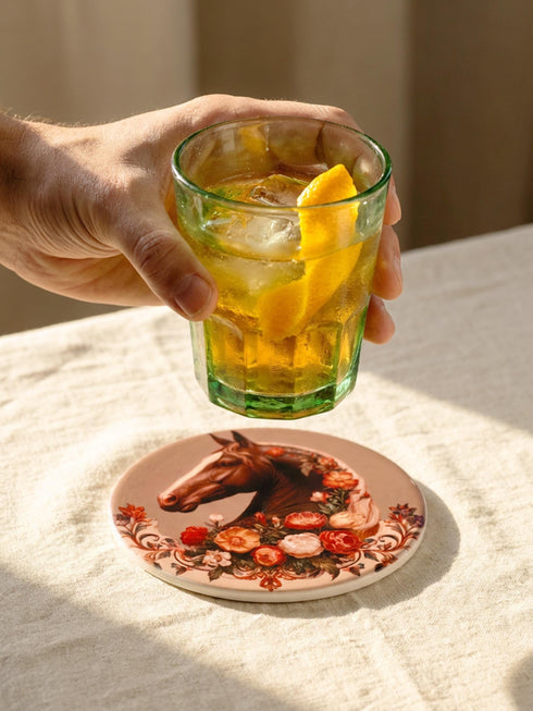 A round glazed ceramic coaster by Grimoire and Goblet featuring a vintage Victorian horse portrait design. The coaster has a high gloss porcelain-like finish and a natural cork bottom for furniture protection. The artwork depicts a dark bay horse head in profile surrounded by moody botanical flowers in shades of rust and deep red, evoking a dark academia and gothic cottagecore aesthetic. This 4x4 inch tile serves as a protective drink mat or decorative equine art accent.