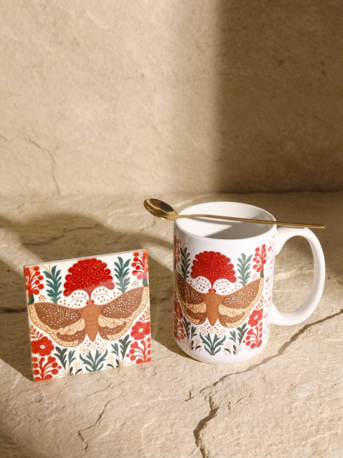 A detailed close-up of a luxury ceramic mug and matching square coaster set from Grimoire and Goblet. The items feature a vintage-style scientific illustration of a large moth with intricate wing patterns in earth tones of rust red and moss green, resting on stylized botanical foliage. The aesthetic is Dark Academia and Victorian Gothic. The mug is a large 15oz white ceramic vessel with a sturdy C-handle. The coaster is a glazed ceramic tile with a glossy finish.