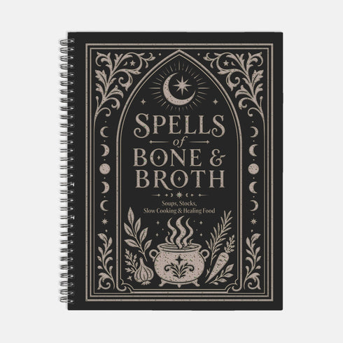 Spiral bound hardcover recipe journal lying flat. The matte black front cover is illustrated with light beige botanical vines, small crescent moons, a radial starburst, and a central cauldron releasing curls of steam, with classical lettering reading Spells of Bone and Broth. A black metal wire-o coil binds the left edge. The interior shows bright white pages printed with a structured recipe template, including labeled sections for ingredients and instructions, over a rigid chipboard backing.