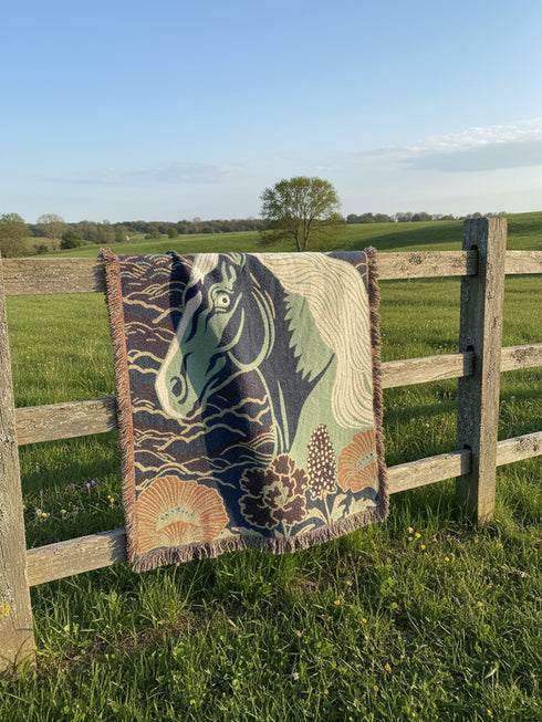 A Grimoire and Goblet woven cotton blanket featuring a large, stylized horse head in sage green and teal against a deep midnight navy background with wave patterns. The bottom edge features woven floral motifs in rust orange and deep plum. The design is a jacquard weave, not printed, with visible thread texture and multi-colored fringe edges. Chinoiserie and Art Nouveau style equestrian tapestry throw.