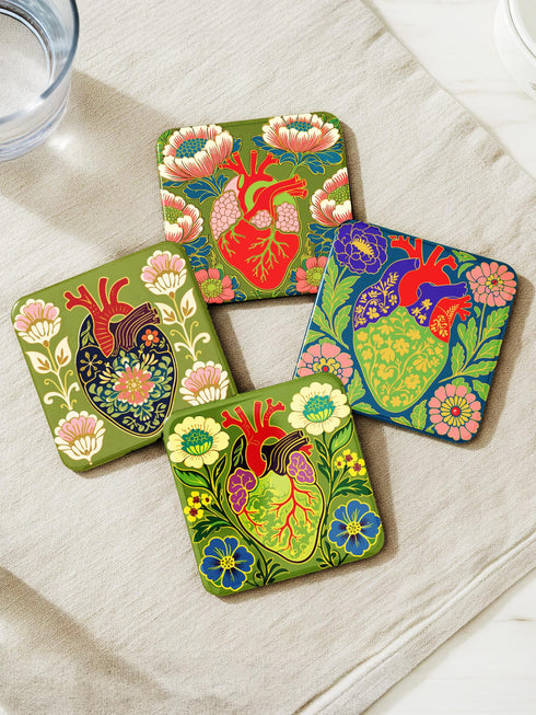 A set of four premium 4x4 inch anatomical heart coasters from Grimoire and Goblet. Each square coaster is made of masonite hardboard with a high-gloss finish and a non-slip cork base. The designs feature intricate biological heart illustrations with folk-art floral motifs in vibrant crimson, pink, yellow, and purple against mossy green and deep teal backgrounds. The high-fidelity print mimics a luxury embroidered texture, perfect for a cardiology gift or dark academia home decor.