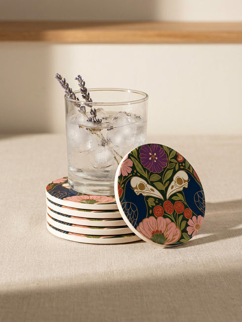 A square drink coaster with a high-gloss finish and natural cork backing. The centralized, symmetrical design features two detailed, cream-colored avian bird skulls facing inward. They are framed by lush botanical flora, including a large purple flower, blush pink blooms, rust-red roses, and green ferns, all set against a deep navy blue background with gold linework accents. Functional gothic, whimsigoth, and dark academia drinkware designed by the Grimoire and Goblet brand.
