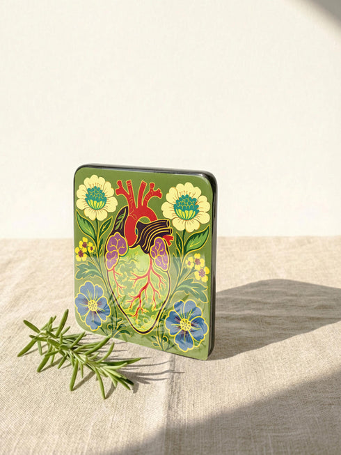 A set of four premium 4x4 inch anatomical heart coasters from Grimoire and Goblet. Each square coaster is made of masonite hardboard with a high-gloss finish and a non-slip cork base. The designs feature intricate biological heart illustrations with folk-art floral motifs in vibrant crimson, pink, yellow, and purple against mossy green and deep teal backgrounds. The high-fidelity print mimics a luxury embroidered texture, perfect for a cardiology gift or dark academia home decor.