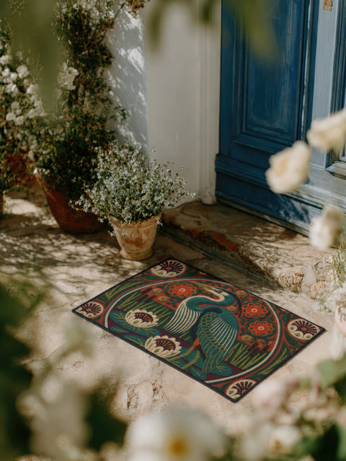 Rectangular low profile doormat printed with a faux embroidery effect that mimics raised crewel stitching. A tall teal blue heron stands at the center among olive green reeds, cattails, and cream water lilies. The rust orange ground is filled with curving botanical vines and small floral medallions, framed by a thin circular border with decorative sunburst corners. A sleek solid black rubber trim finishes all four edges of the one sided print.