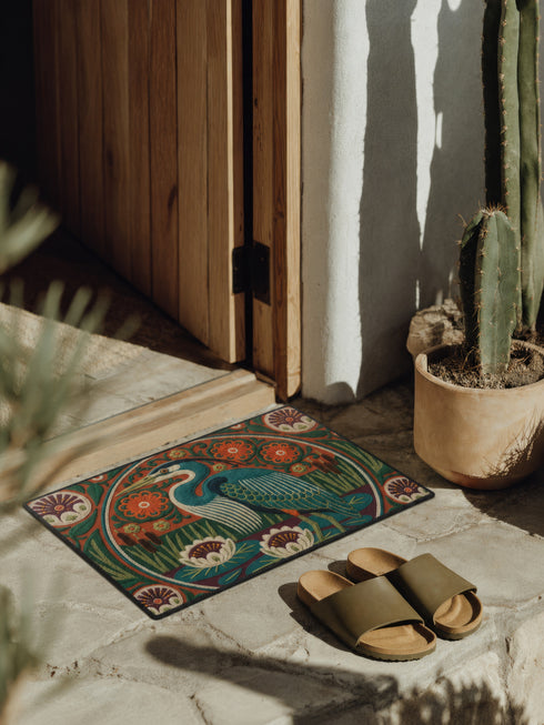 Rectangular low profile doormat printed with a faux embroidery effect that mimics raised crewel stitching. A tall teal blue heron stands at the center among olive green reeds, cattails, and cream water lilies. The rust orange ground is filled with curving botanical vines and small floral medallions, framed by a thin circular border with decorative sunburst corners. A sleek solid black rubber trim finishes all four edges of the one sided print.