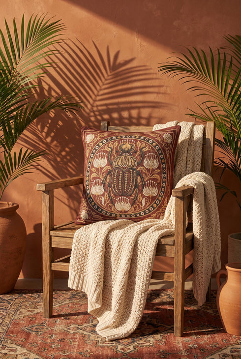 Square 17 by 18 inch jacquard woven throw pillow, woven identically on both faces and closed with a clean finished edge. A large detailed scarab beetle in black, gold, and dark brown sits at the center inside a dotted circular border, surrounded by symmetrical Art Nouveau vines and cream lotus flowers on a rust red ground. Small white floral motifs sit in each corner. The design is woven flat into the cotton blend cloth, with visible thread texture and a soft sheen in the ground.