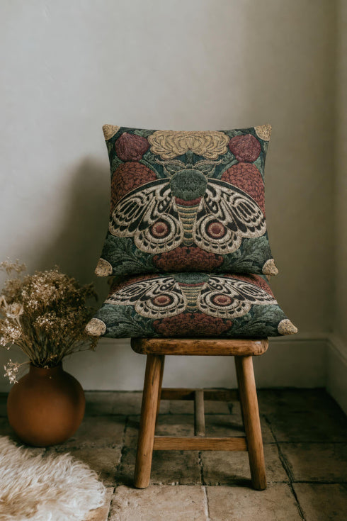 Square 17 by 18 inch jacquard woven throw pillow, woven identically on both faces and closed with a clean sewn edge. A symmetrical emperor moth with wings detailed in black, warm gold, and deep burgundy red spreads across the center of a dark forest green ground. A large golden yellow flower blooms above the moth and burgundy floral buds sit on either side among woven botanical sprays. The design is woven flat into the cotton blend cloth with visible thread texture.