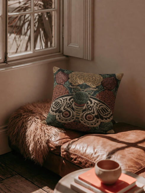 Square 17 by 18 inch jacquard woven throw pillow, woven identically on both faces and closed with a clean sewn edge. A symmetrical emperor moth with wings detailed in black, warm gold, and deep burgundy red spreads across the center of a dark forest green ground. A large golden yellow flower blooms above the moth and burgundy floral buds sit on either side among woven botanical sprays. The design is woven flat into the cotton blend cloth with visible thread texture.