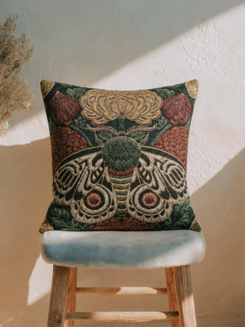 Square 17 by 18 inch jacquard woven throw pillow, woven identically on both faces and closed with a clean sewn edge. A symmetrical emperor moth with wings detailed in black, warm gold, and deep burgundy red spreads across the center of a dark forest green ground. A large golden yellow flower blooms above the moth and burgundy floral buds sit on either side among woven botanical sprays. The design is woven flat into the cotton blend cloth with visible thread texture.